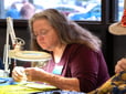 Woman looks through magnified glass light to repair an item. 