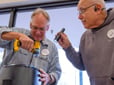 Two men repair an electronic item. One holds a flashlight while the other holds a drill. 